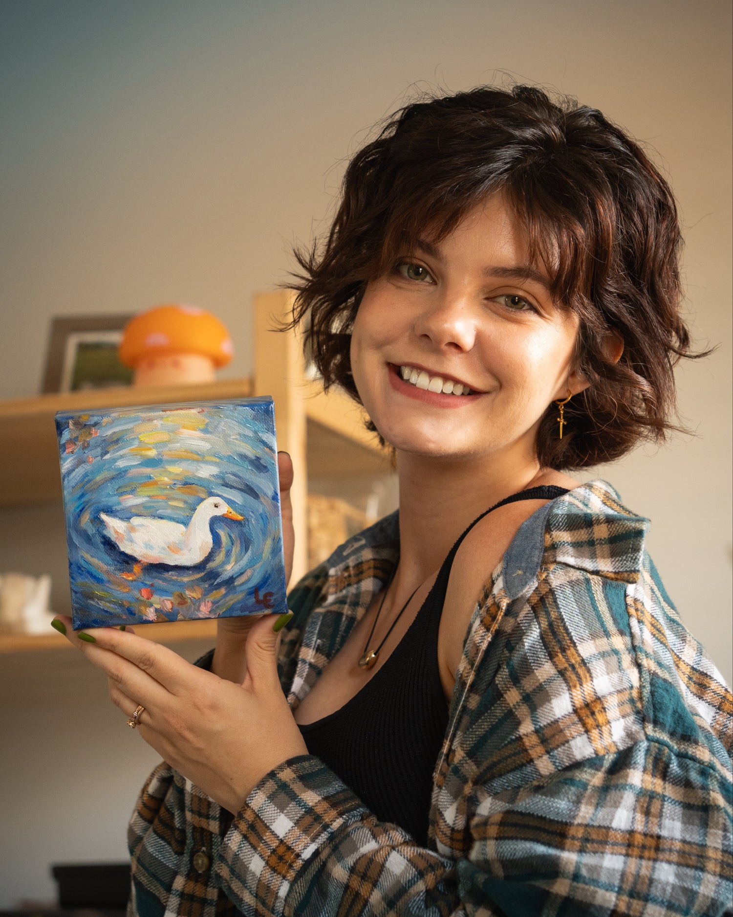 A smiling woman with short dark hair holds up a small square oil painting of a white duck swimming on a blue, impressionist water background. She is wearing an open plaid shirt over a black top in an indoor setting with shelves in the background.